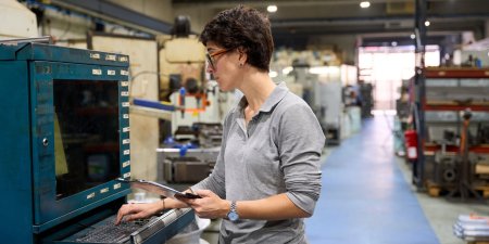 Female engineer using computer and digital tablet, managing industrial machinery in a metallurgy factory producing custom mechanical parts