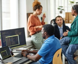 African IT specialist working with software typing on keyboard sitting at table with computers while his colleagues working in team in background