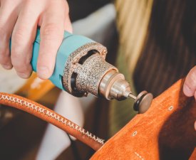 Master manufacturing bag of genuine leather in his workshop at home. Man processes the edge of the product with a grinder machine. Little local business. Macro view.